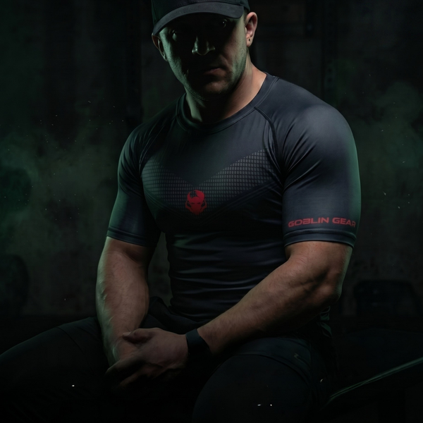 Goblin Gear Redline Compression Tee – man wearing a dark athletic shirt with a logo, sitting in a dark forest