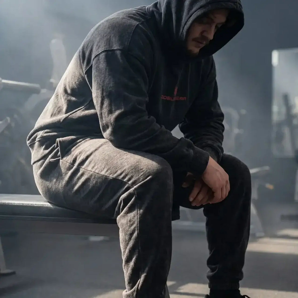 Goblin Gear Acid Wash Hoodie – person wearing a dark hoodie and pants sitting on a bench in a gym
