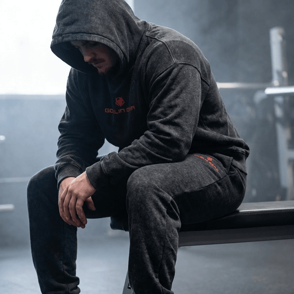 Goblin Gear Acid Wash Hoodie – person wearing a dark hoodie with a logo, sitting on a bench in a blurred indoor setting